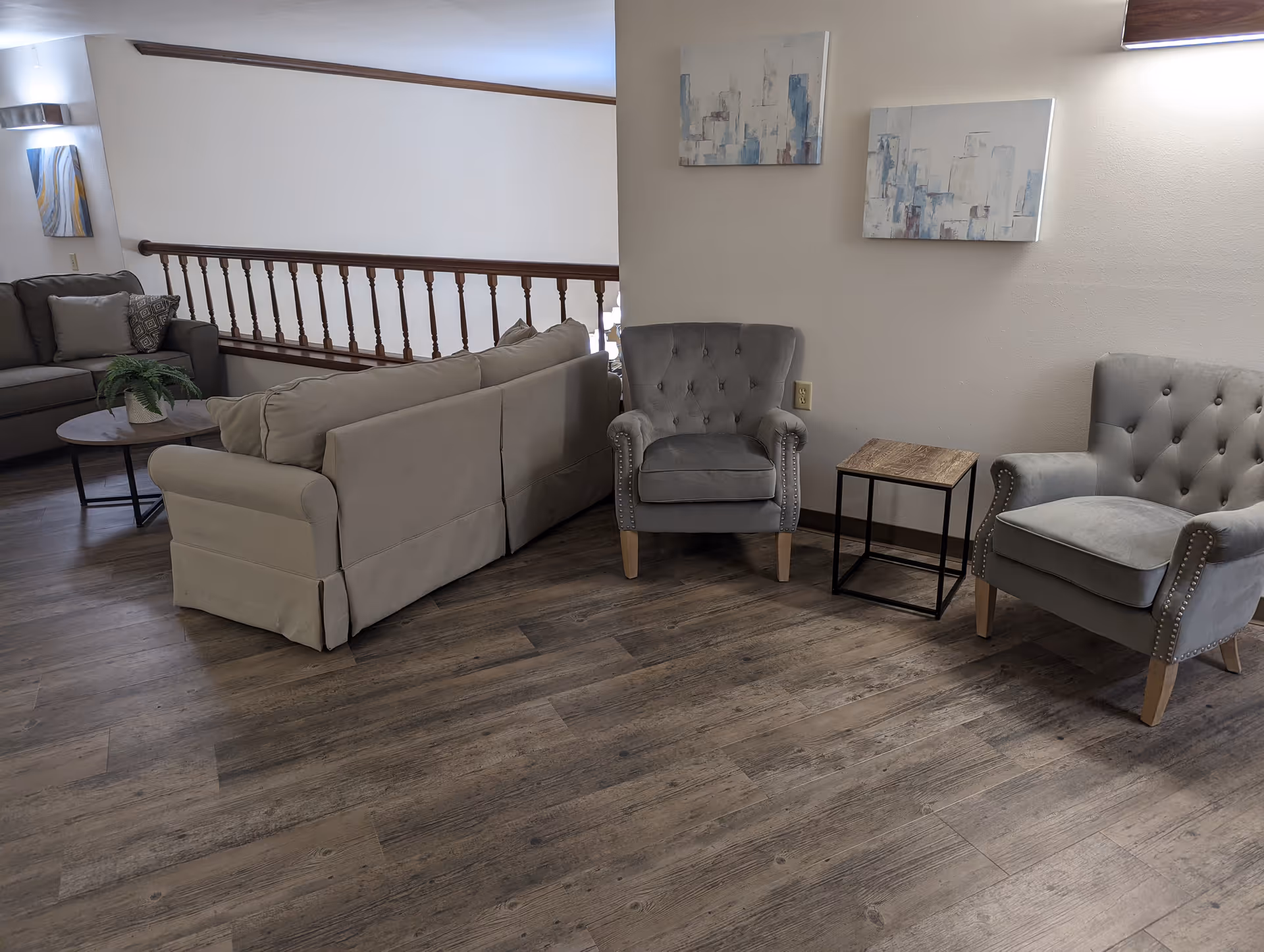 A seating area with a beige sofa, two gray armchairs, a small wooden side table, wall art, and wood-look flooring.