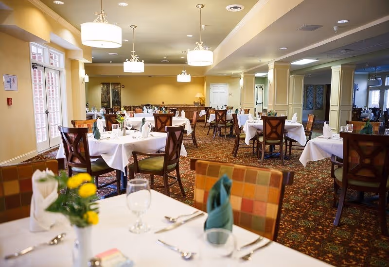 Spacious dining room with multiple tables draped in white linens, place settings, chairs, and hanging pendant lights.