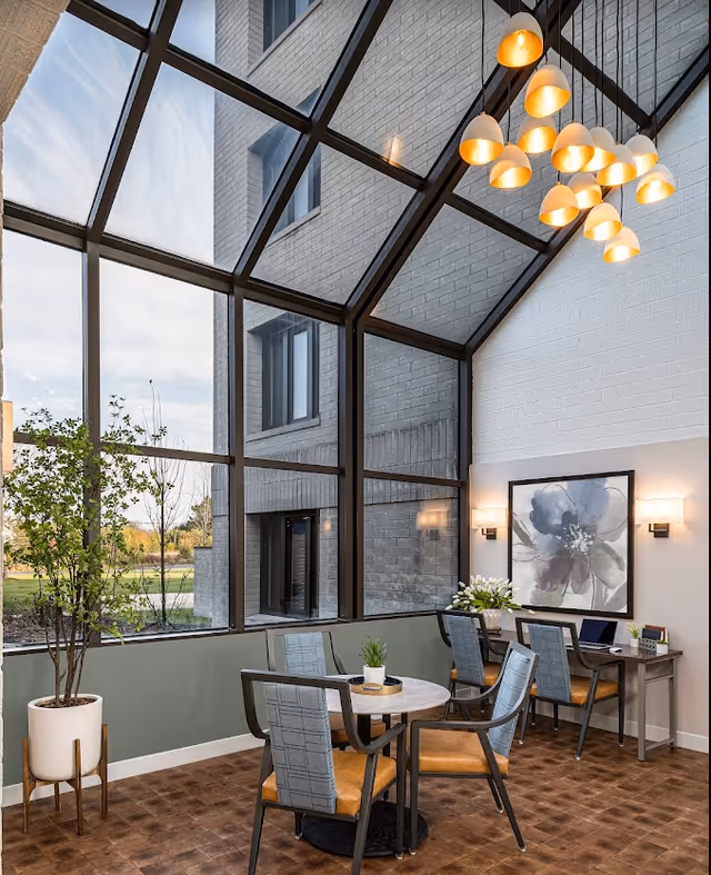 A bright indoor seating area with large floor-to-ceiling windows and a glass ceiling letting in natural light. The space features a round table with four chairs, a potted plant, a desk with two chairs, a large floral artwork on the wall, and a modern chandelier with multiple pendant lights.