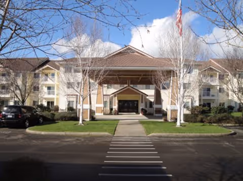 Front exterior view of Country Meadows Village Retirement Community building with a covered entrance, flagpole, leafless trees, parked cars, and a clear sky with clouds.