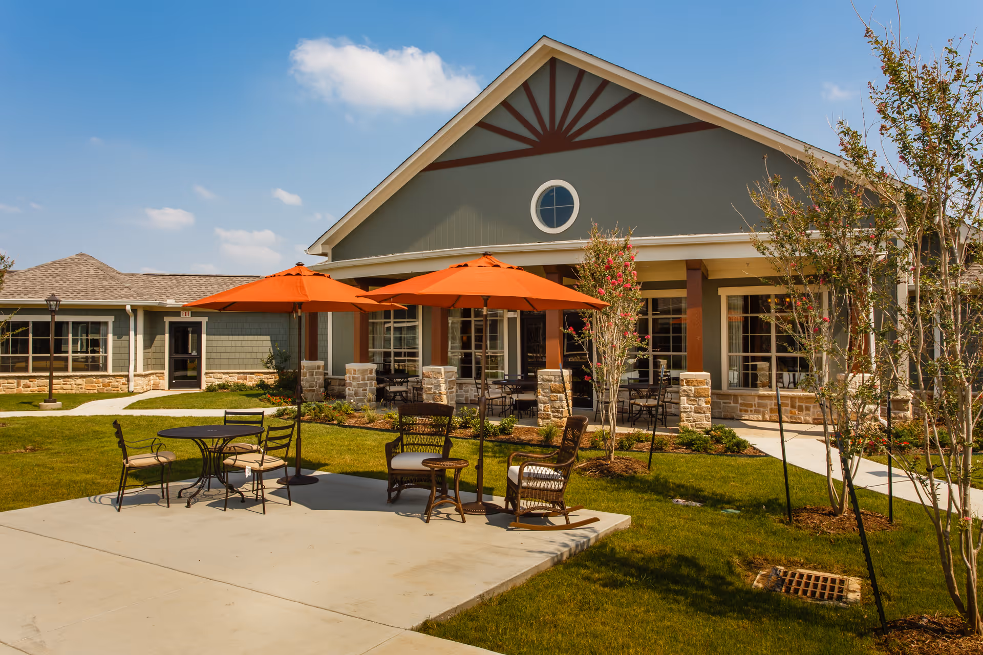 Exterior view of a single-story assisted living building with a patio area featuring orange umbrellas and outdoor seating.