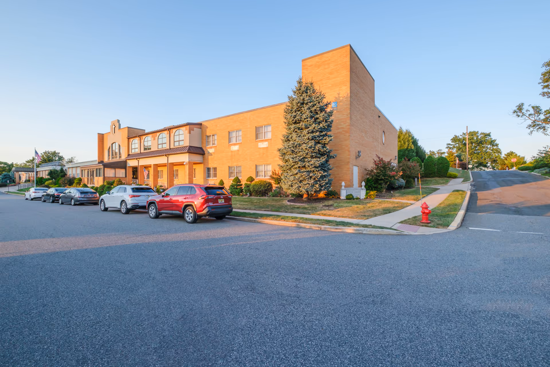 Exterior view of St Joseph's Senior Home - Assisted Living and Nursing Center building during daylight with several cars parked along the street in front. The building is made of light brown brick with multiple windows and a small landscaped area with trees and shrubs near the entrance.