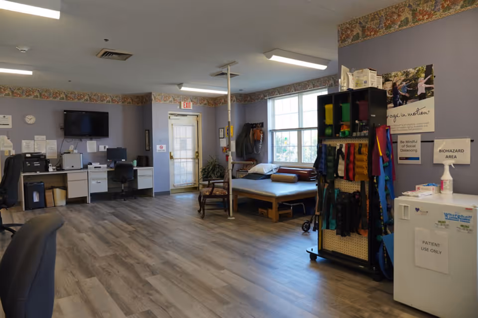 A spacious room with light purple walls and wood flooring, featuring a desk area with computer and printer, a wall-mounted TV, a therapy or examination table near a window, and a rack holding various exercise or therapy equipment. Signs on the wall indicate a biohazard area and reminders for social distancing.