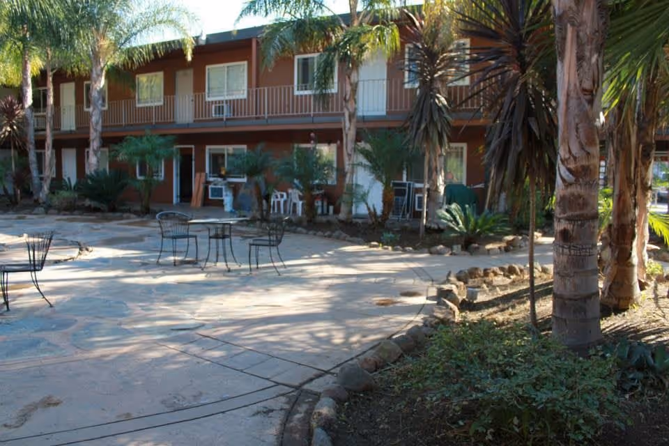 Outdoor patio area with metal tables and chairs surrounded by palm trees and plants, adjacent to a two-story building with balconies and windows.