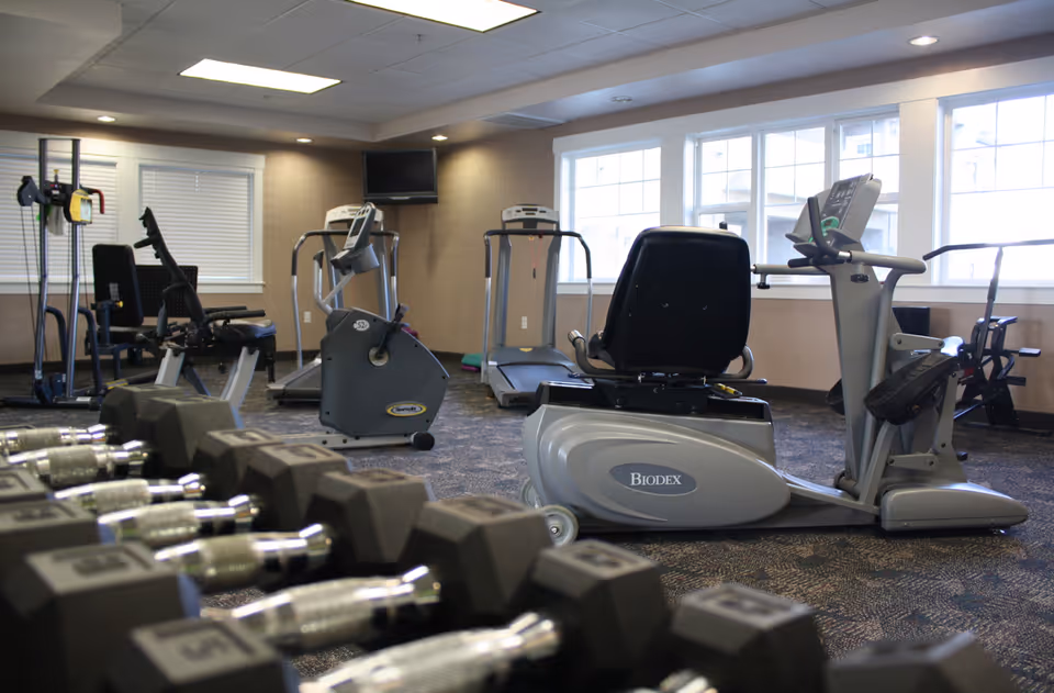 Interior fitness room with cardio machines, treadmills, and a row of dumbbells in front of windows.