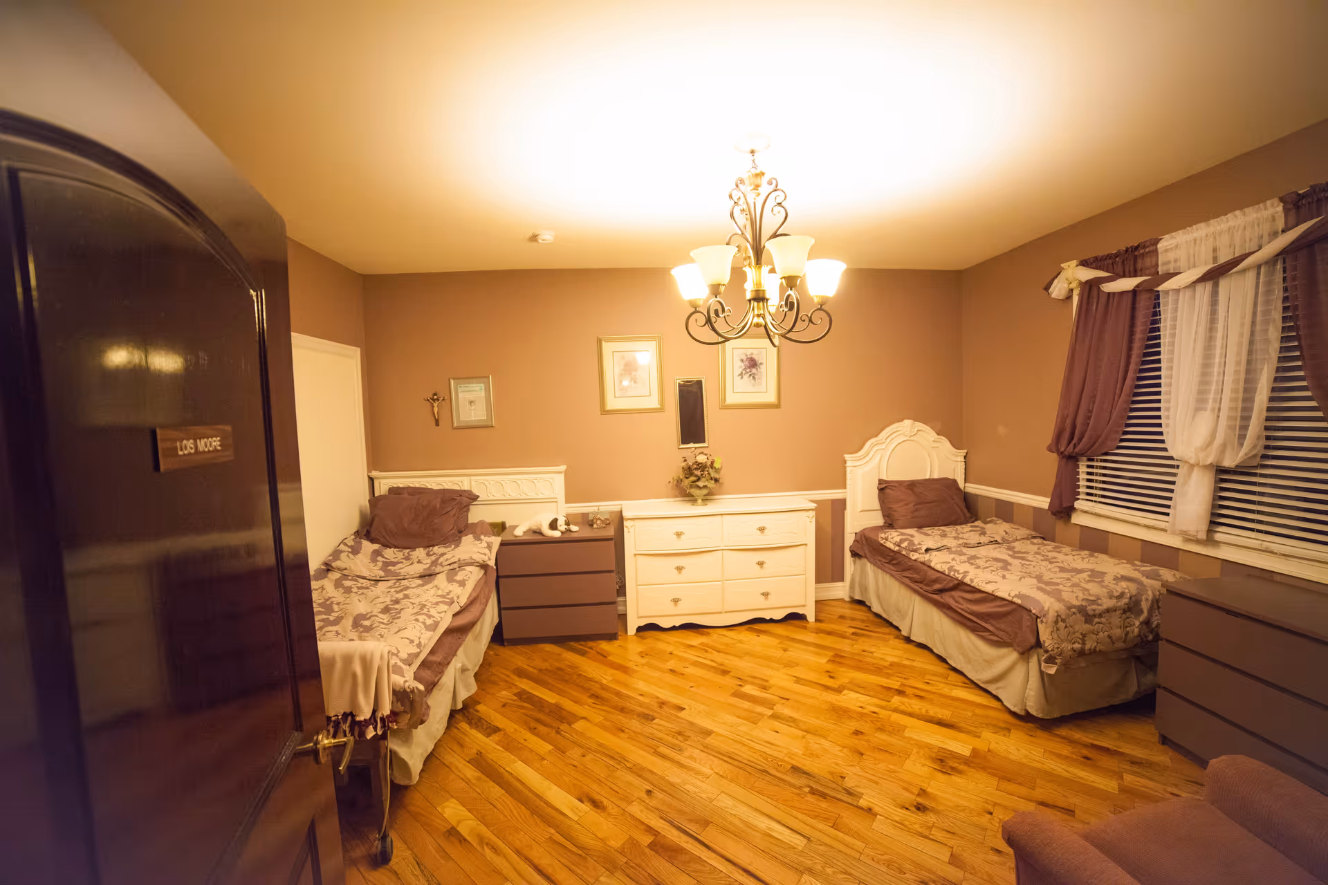 A warmly lit bedroom with two single beds, each with brown and beige bedding. Between the beds is a white dresser with a small mirror and decorative items on top. There are two framed pictures on the wall above the dresser. The room has wooden flooring, a chandelier light fixture, and a window with brown and white curtains. A door with a nameplate reading 'LOS MOORE' is partially open on the left side of the image.