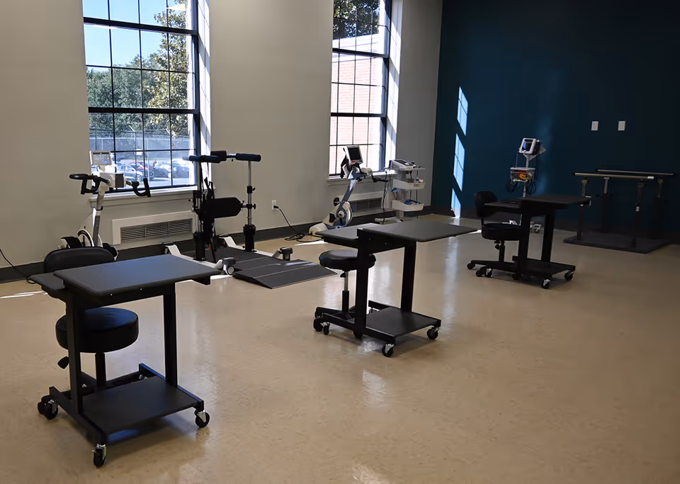 A rehabilitation and wellness room with exercise equipment including stationary bikes, therapy tables with stools on wheels, and medical monitoring devices. The room has large windows letting in natural light and a blue accent wall.