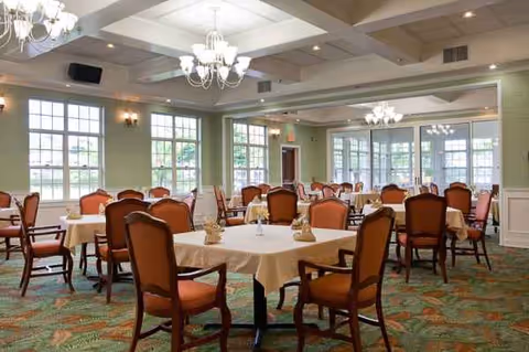 Spacious dining room with round tables, orange chairs, and chandeliers in a senior care facility.