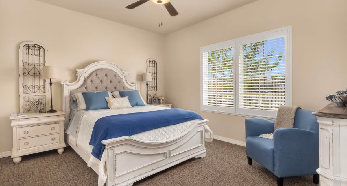 A cozy bedroom with a white wooden bed frame featuring a tufted headboard, dressed in white and blue bedding. There are two matching white nightstands with lamps on either side of the bed. A blue upholstered armchair with a beige throw blanket is positioned near a window with white blinds, letting in natural light. The walls are painted beige and the floor is carpeted.