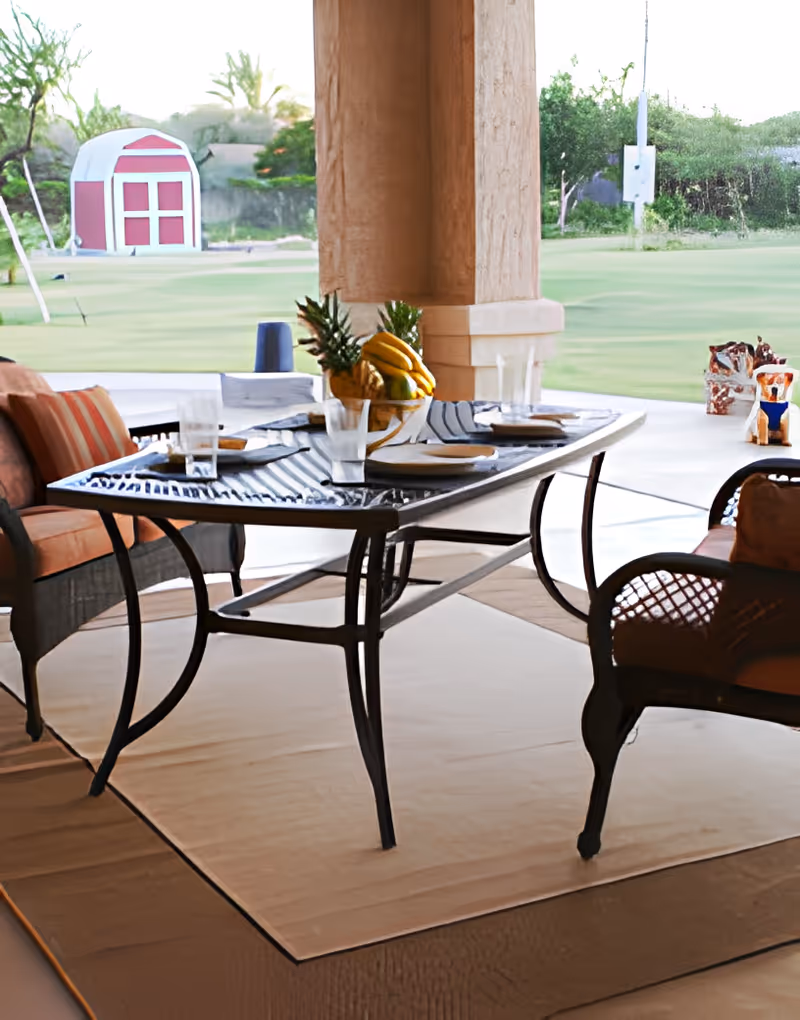 Outdoor patio dining area with a glass-top table set with plates, glasses, and a centerpiece of bananas and pineapples. The table is surrounded by cushioned chairs and overlooks a green lawn with a red and white shed in the background.