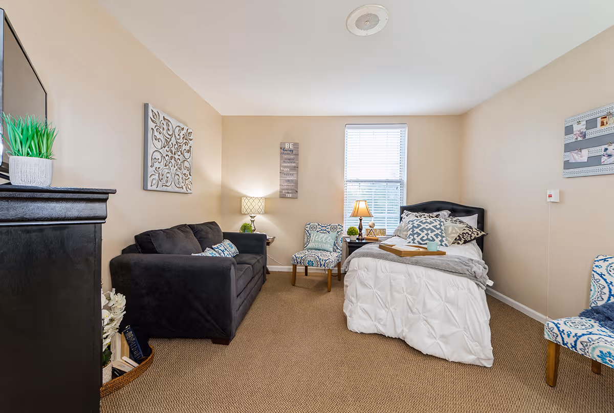 Furnished bedroom with a twin bed, black sofa, patterned chairs, a dresser-mounted TV and a window.
