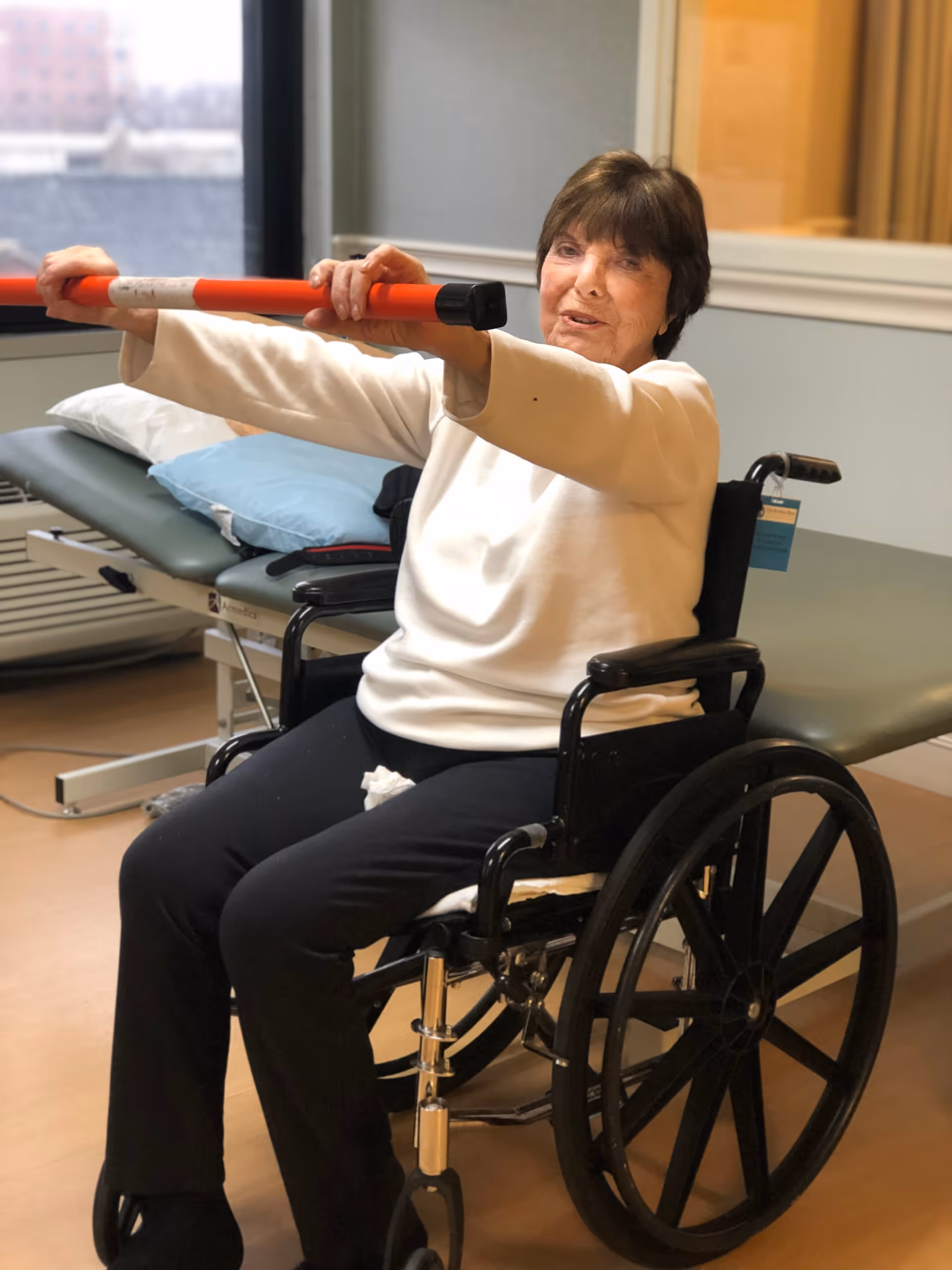 An elderly woman sitting in a wheelchair indoors, holding a red and white exercise bar with both hands stretched out in front of her. Behind her is a padded examination or treatment table with pillows, and a large window showing an urban view outside.