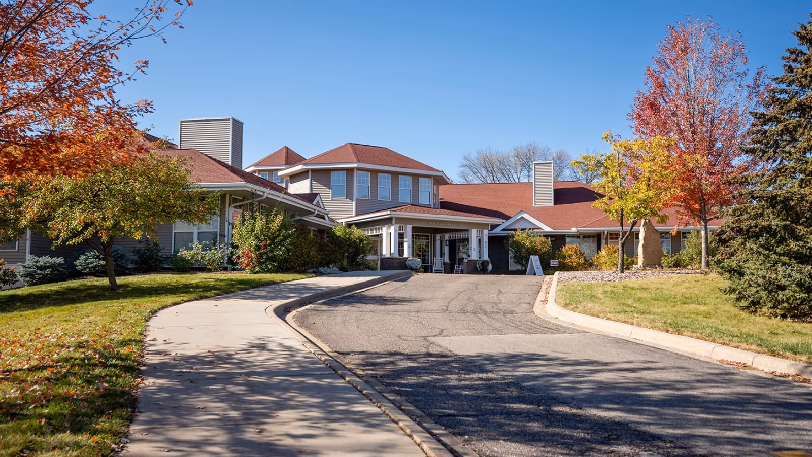 Front exterior view of Three Links Care Center building with a driveway and sidewalk leading to the entrance, surrounded by green grass and trees with autumn foliage under a clear blue sky.