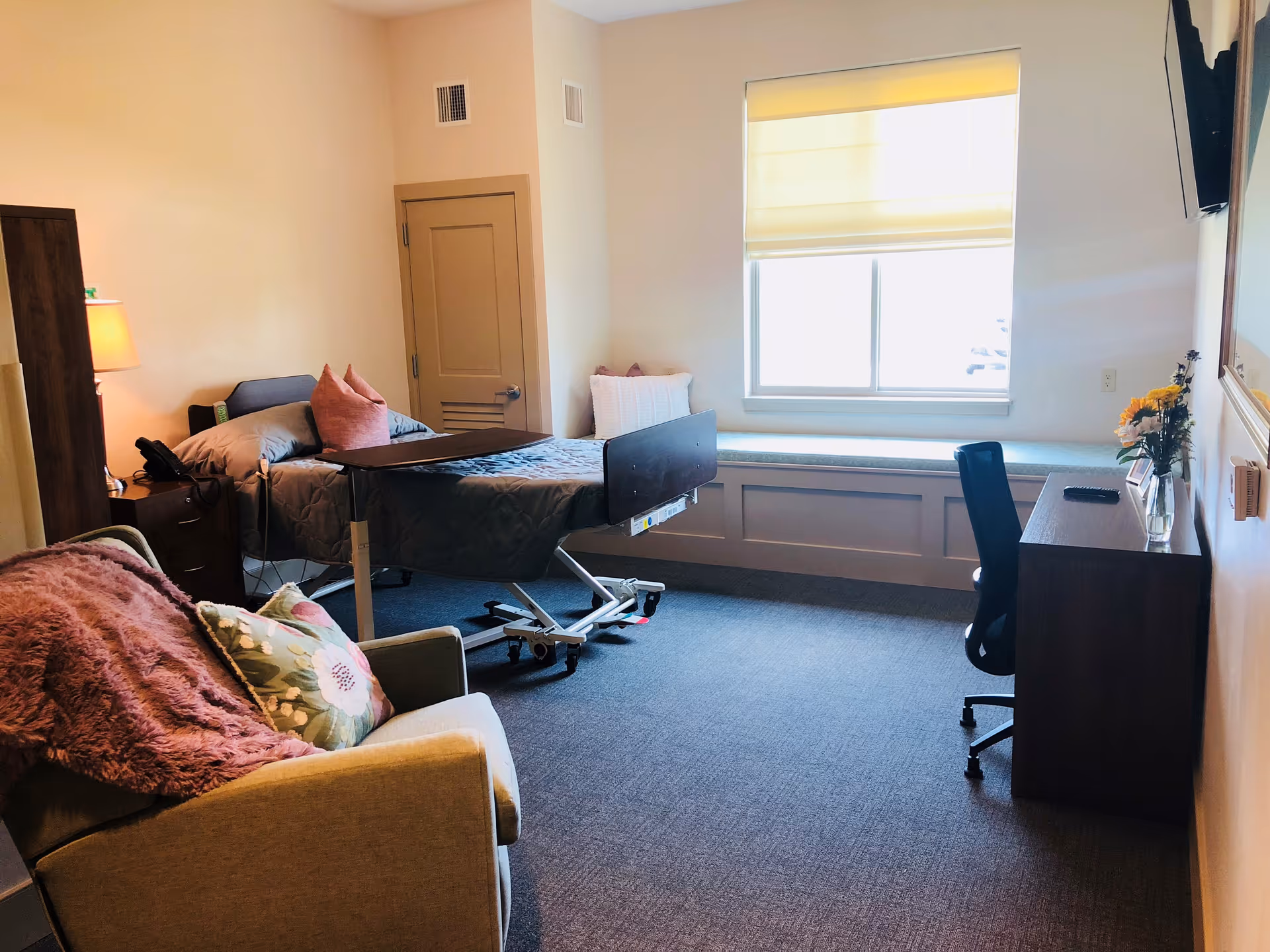 Well-lit resident bedroom with an adjustable hospital bed, armchair, desk and a window seat.