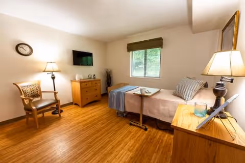 A senior living facility bedroom with a single bed covered with a beige blanket and a blue throw. There is a wooden dresser with a flat-screen TV mounted above it, a wooden chair with a cushion, a small table on wheels next to the bed, and a wooden nightstand with a lamp and a glass of water. A window with a green valance lets in natural light, and the floor is made of wood.