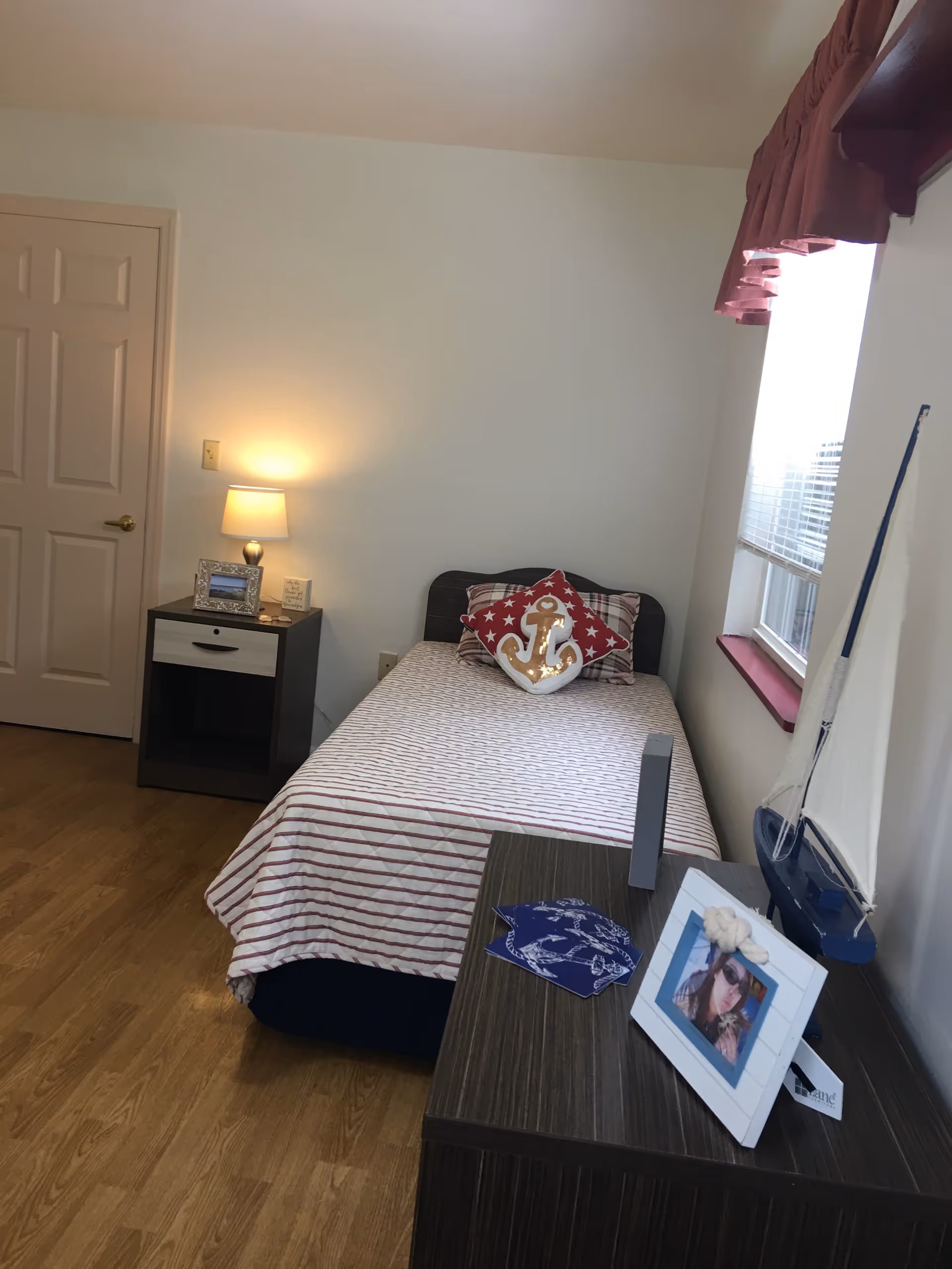 Small bedroom with a single bed against the wall, a nightstand with a lamp, and a dresser holding a framed photo and decorative sailboat by the window.