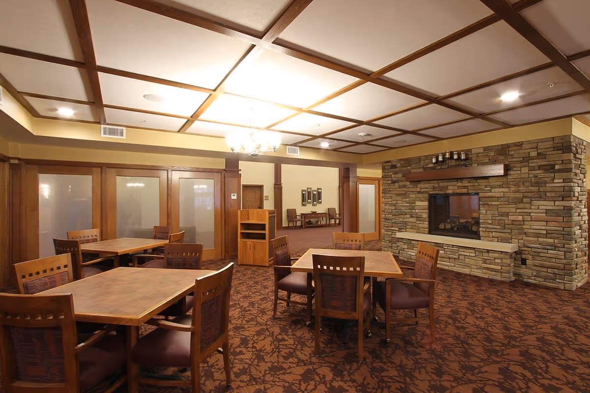 Interior view of a senior living facility common area with wooden tables and chairs arranged on a patterned carpet. The room features a stone fireplace on the right wall, wooden paneling, and a coffered ceiling with recessed lighting and a chandelier. In the background, there are additional seating areas and wooden doors with frosted glass panels.