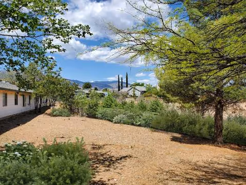 Outdoor view of a landscaped area with trees, bushes, and a dirt ground at Christian Care Cottonwood facility. Buildings are visible on the left side, with a clear blue sky and mountains in the background.