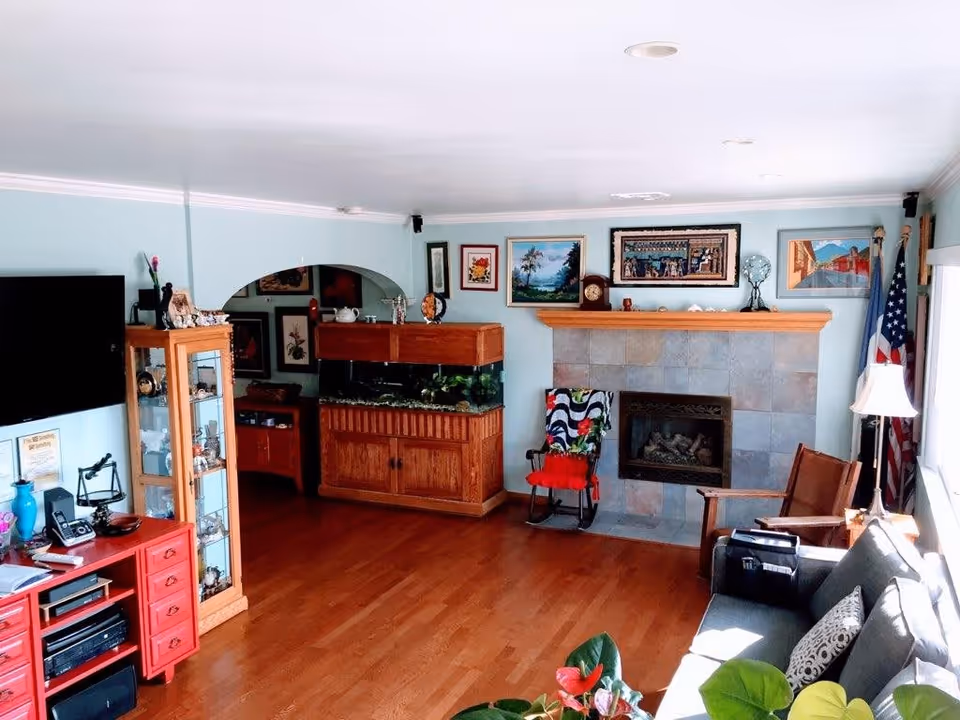 A cozy living room with light blue walls, wooden flooring, and a tiled fireplace with a wooden mantel. The room features a gray sofa with patterned cushions, a wooden rocking chair with a colorful blanket, a wooden cabinet with a fish tank on top, and a red media console with electronic devices. Various framed paintings and decorations adorn the walls, and an American flag stands near a window with a white lamp beside it.