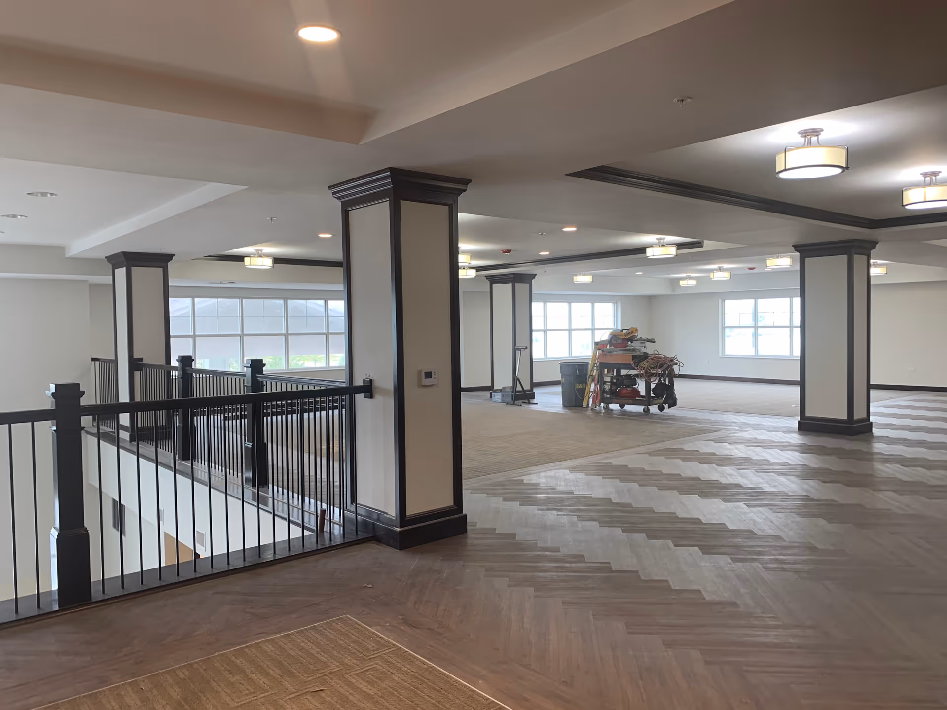 Spacious interior area with large windows, multiple square columns with dark trim, and a railing overlooking a lower level. The floor features a combination of carpet and wood in a herringbone pattern. Ceiling lights are evenly spaced, and there is a cart with cleaning or maintenance supplies in the background.