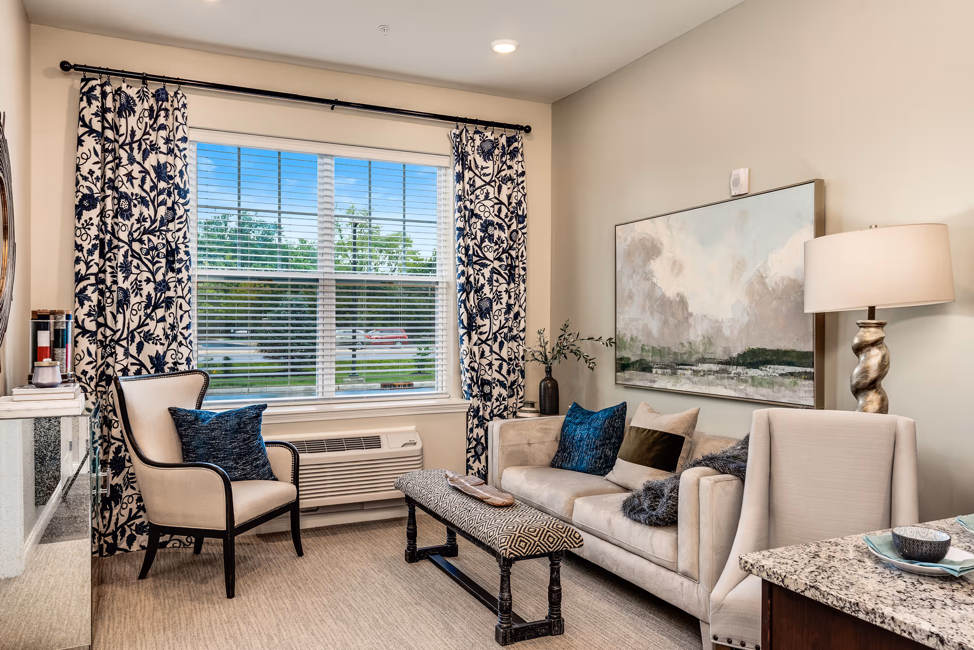 A cozy living room area with a beige sofa adorned with blue and brown pillows, a patterned bench in front of the sofa, a beige armchair with a blue pillow, and a large window with floral curtains letting in natural light. A table lamp with a twisted base and a large abstract painting hang on the wall behind the sofa.