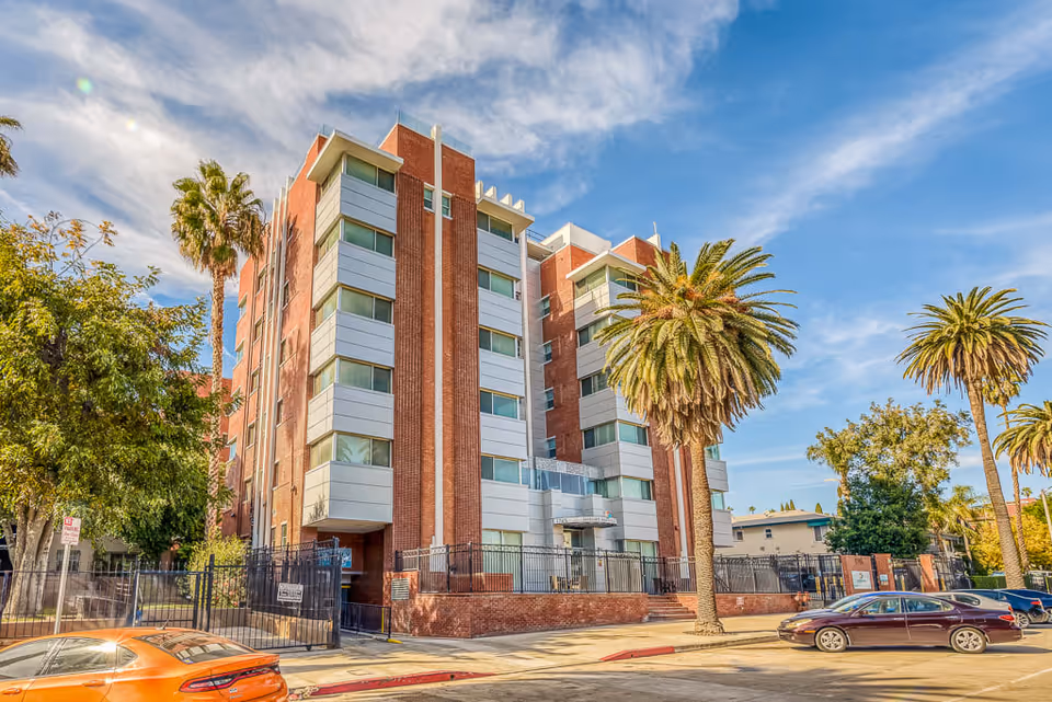 Exterior view of a multi-story senior living facility building with red brick and white paneling, surrounded by palm trees and other greenery under a partly cloudy blue sky. Several cars are parked along the street in front of the building.