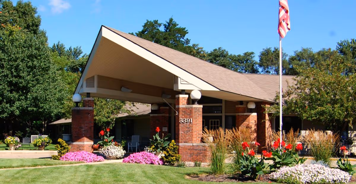 Front entrance of a low brick building with a covered portico, manicured lawn, colorful flower beds and an American flag.