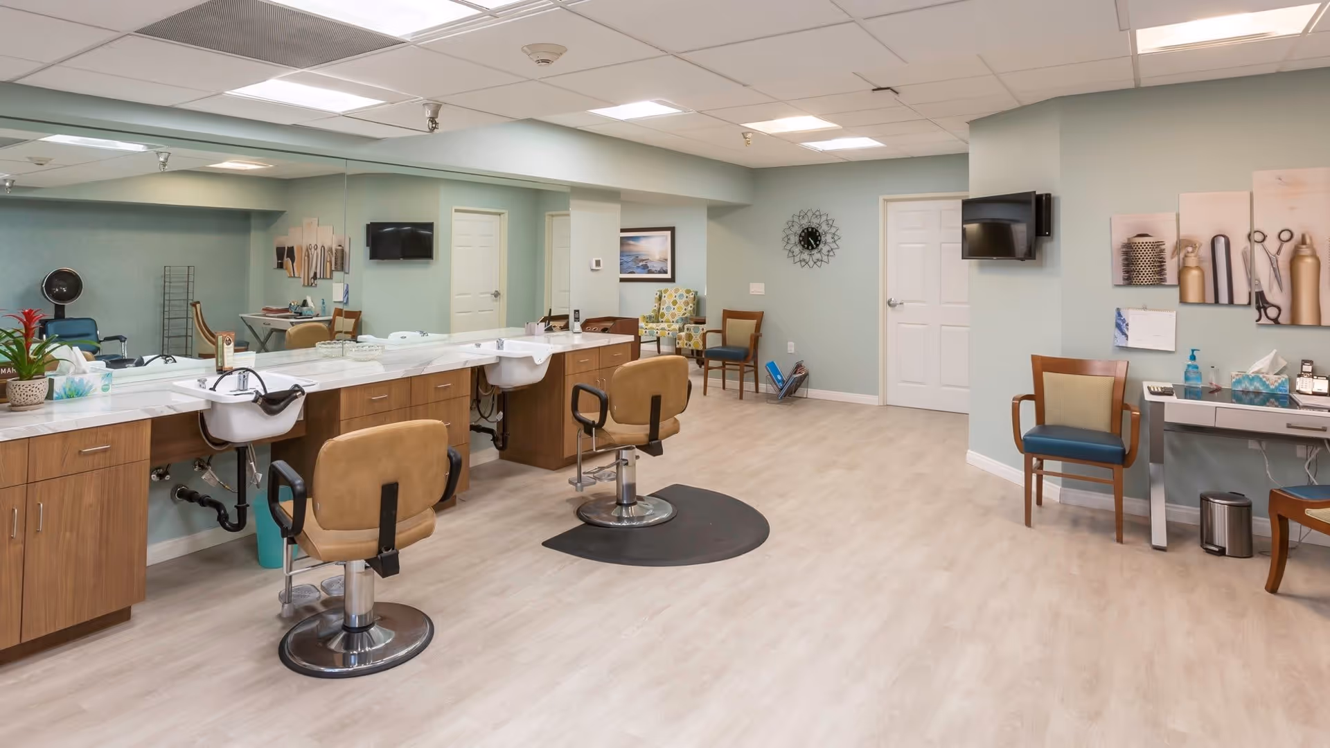 Interior view of a salon area in a senior living facility named Royal Oaks, featuring two salon chairs with sinks, a large mirror along the wall, wooden cabinetry, a small seating area with chairs, a wall-mounted TV, and hair styling decor on the walls.