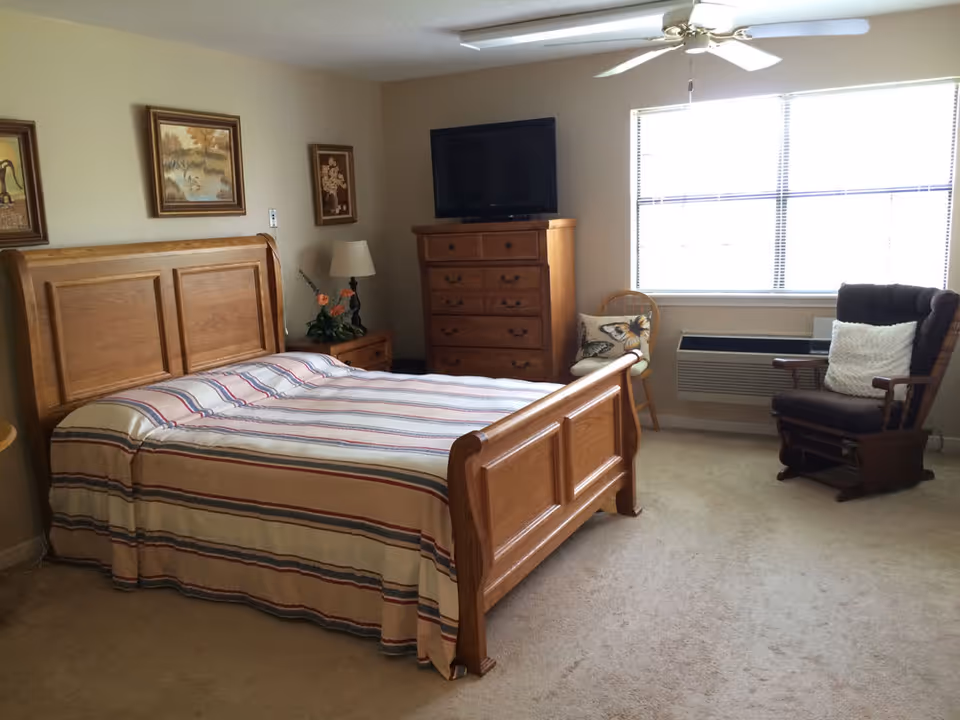 A bedroom with a wooden bed frame and striped bedspread, a wooden dresser with a TV on top, a nightstand with a lamp and flowers, a wooden chair with a butterfly cushion, and a cushioned rocking chair near a window with blinds.
