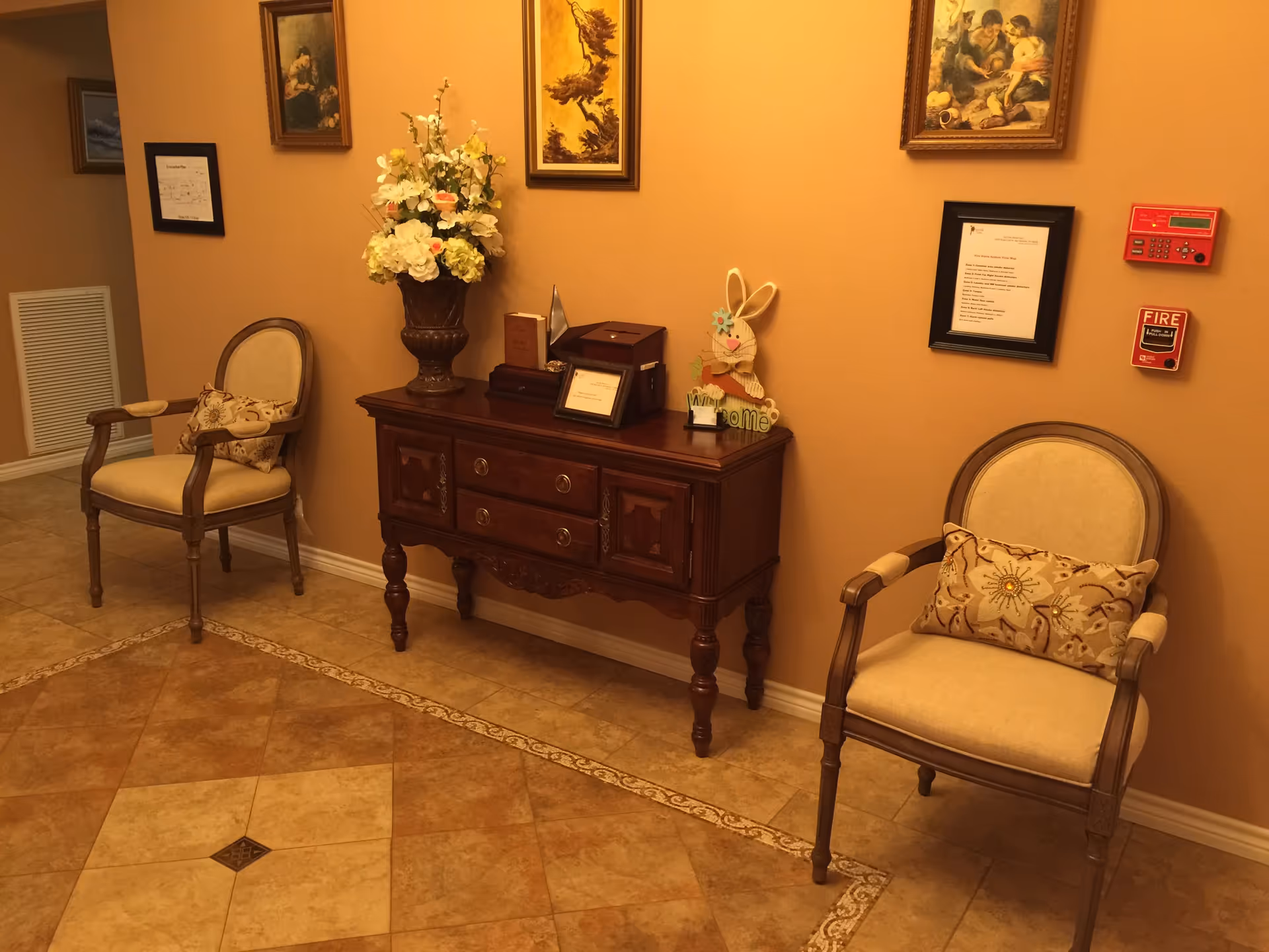A cozy interior hallway with two beige upholstered chairs with decorative pillows on either side of a wooden console table. The table holds a large vase with white and light pink flowers, a wooden bunny decoration with a 'Welcome' sign, and framed pictures. The walls are painted a warm beige color and adorned with framed artwork and a fire alarm system.