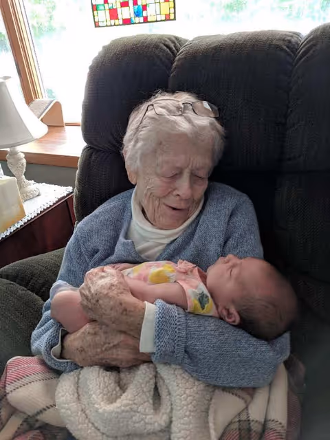 An elderly woman with glasses resting on her head is sitting in a cushioned chair, gently holding and looking lovingly at a sleeping baby wrapped in a blanket.