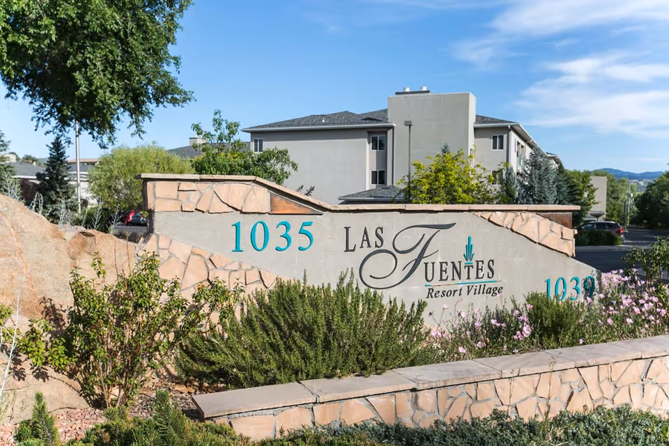 Stone and stucco sign for Las Fuentes Resort Village with address numbers 1035 and 1030, surrounded by greenery and flowers, with a multi-story building and trees in the background under a blue sky.