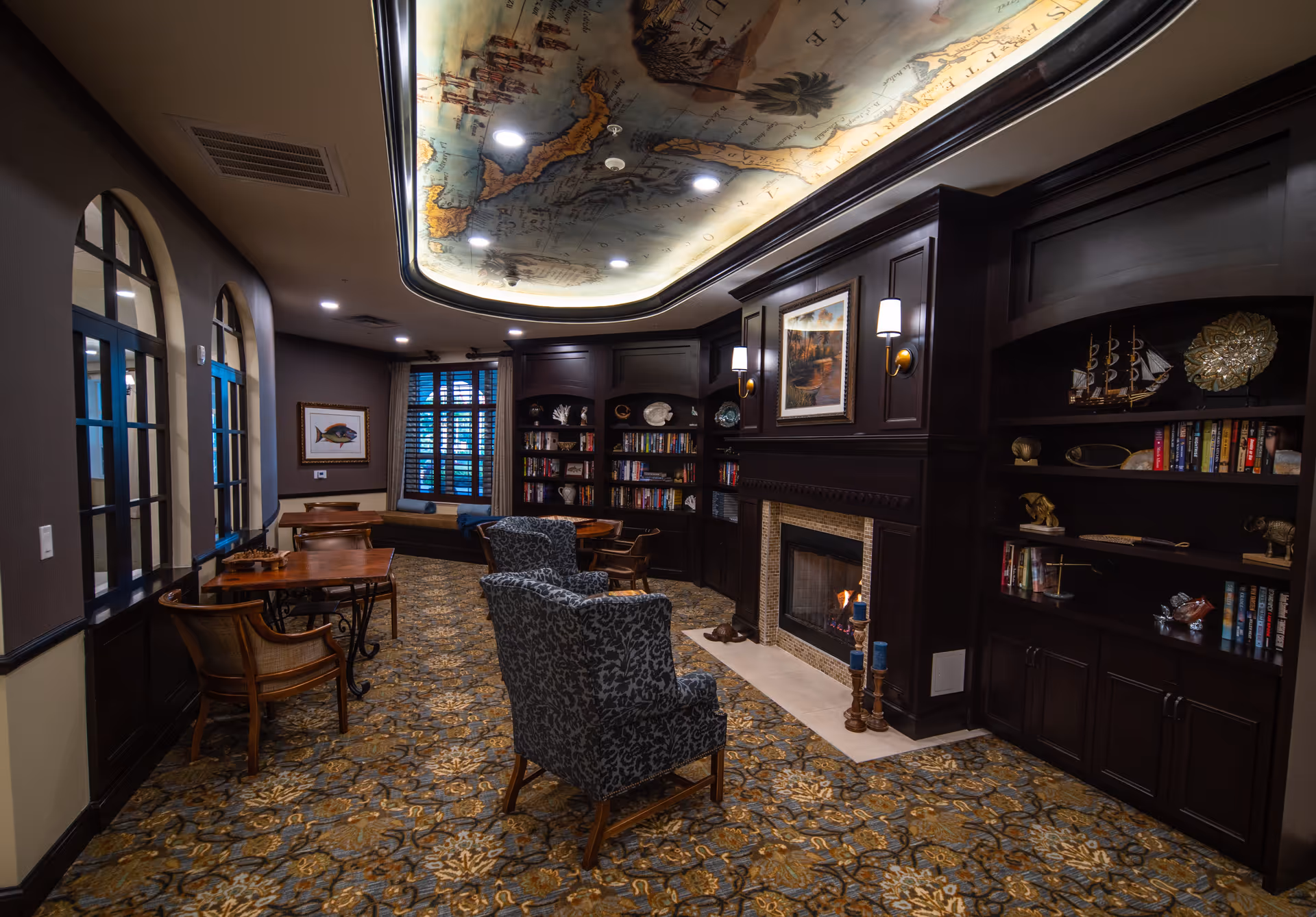 Cozy common room with a lit fireplace, built-in dark wood bookshelves, patterned carpet, seating and a decorative map-painted ceiling.