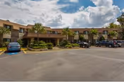 Exterior view of a two-story senior living facility building with a parking lot in front. Several cars are parked, and there are palm trees and landscaped greenery around the building under a partly cloudy sky.