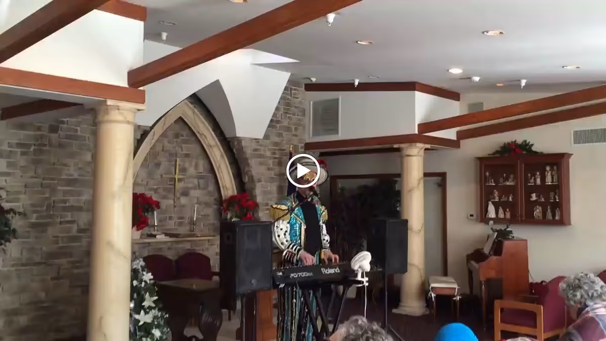 An indoor common area with stone walls and decorative columns, featuring a person dressed in a colorful costume playing a keyboard. There are chairs and a piano in the background, along with shelves displaying figurines and plants.