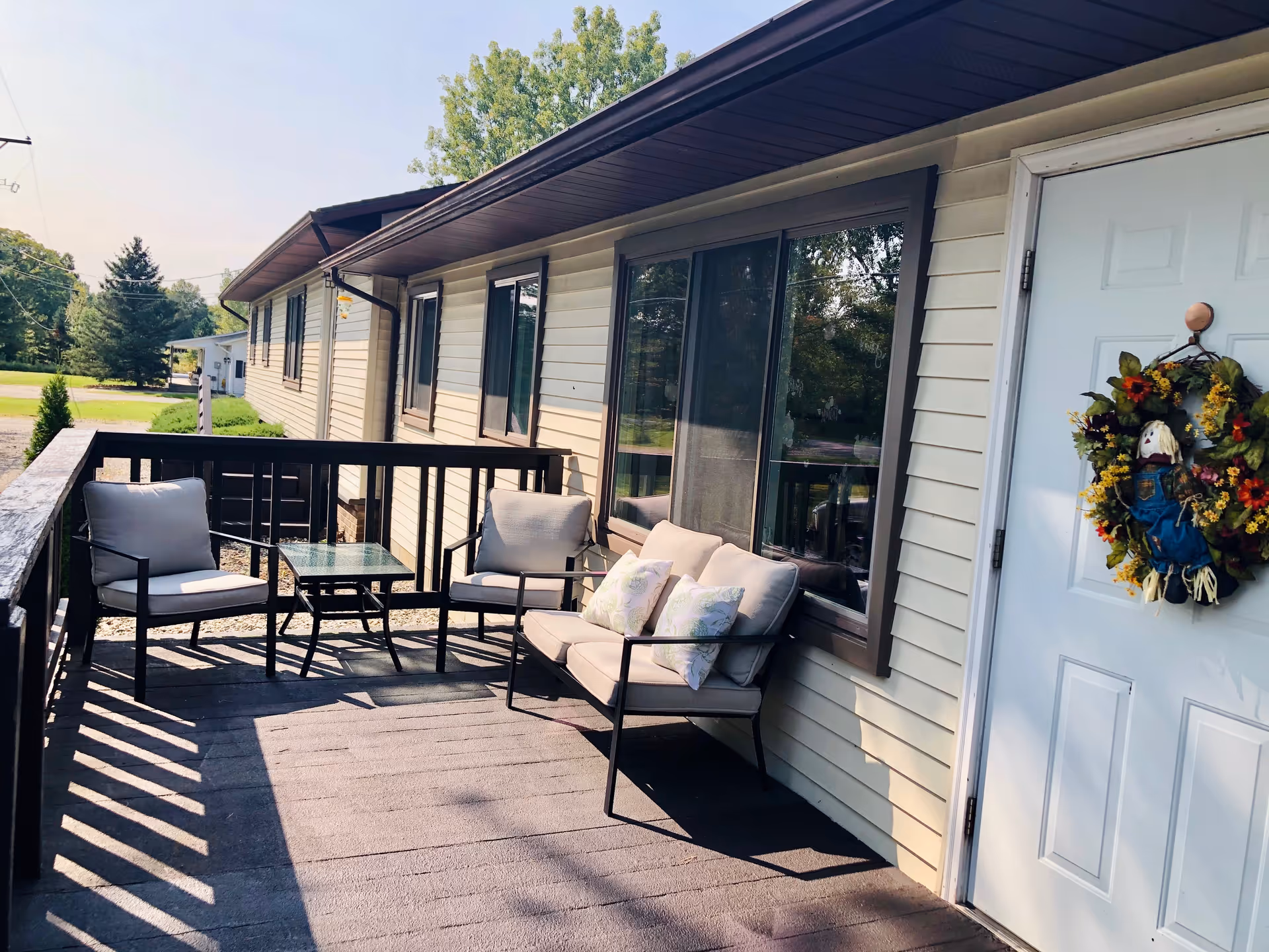 Outdoor patio area with cushioned chairs and a small glass table, attached to a beige building with a white door decorated with a colorful wreath. Trees and greenery are visible in the background.