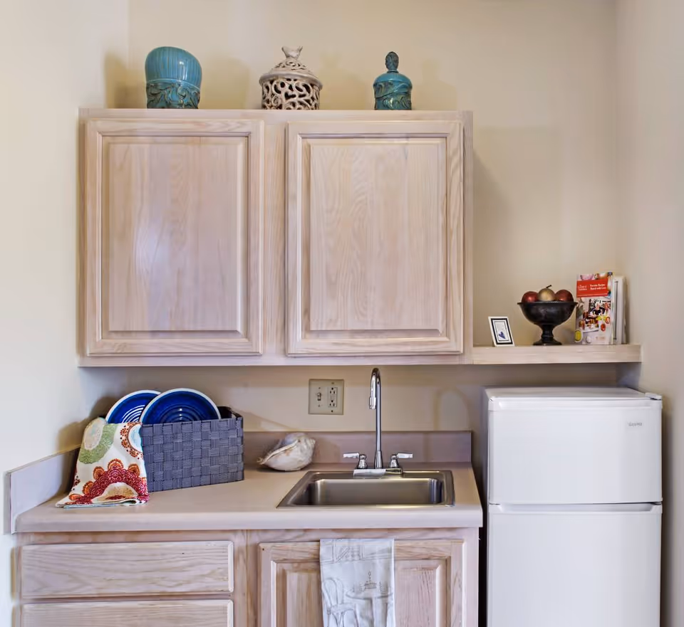 Small kitchenette area with light wood cabinets, a stainless steel sink with a faucet, a small white refrigerator, and a countertop holding a basket with plates, a decorative shell, and a bowl of fruit. There are decorative jars on top of the upper cabinets and a small shelf with brochures and a framed picture.