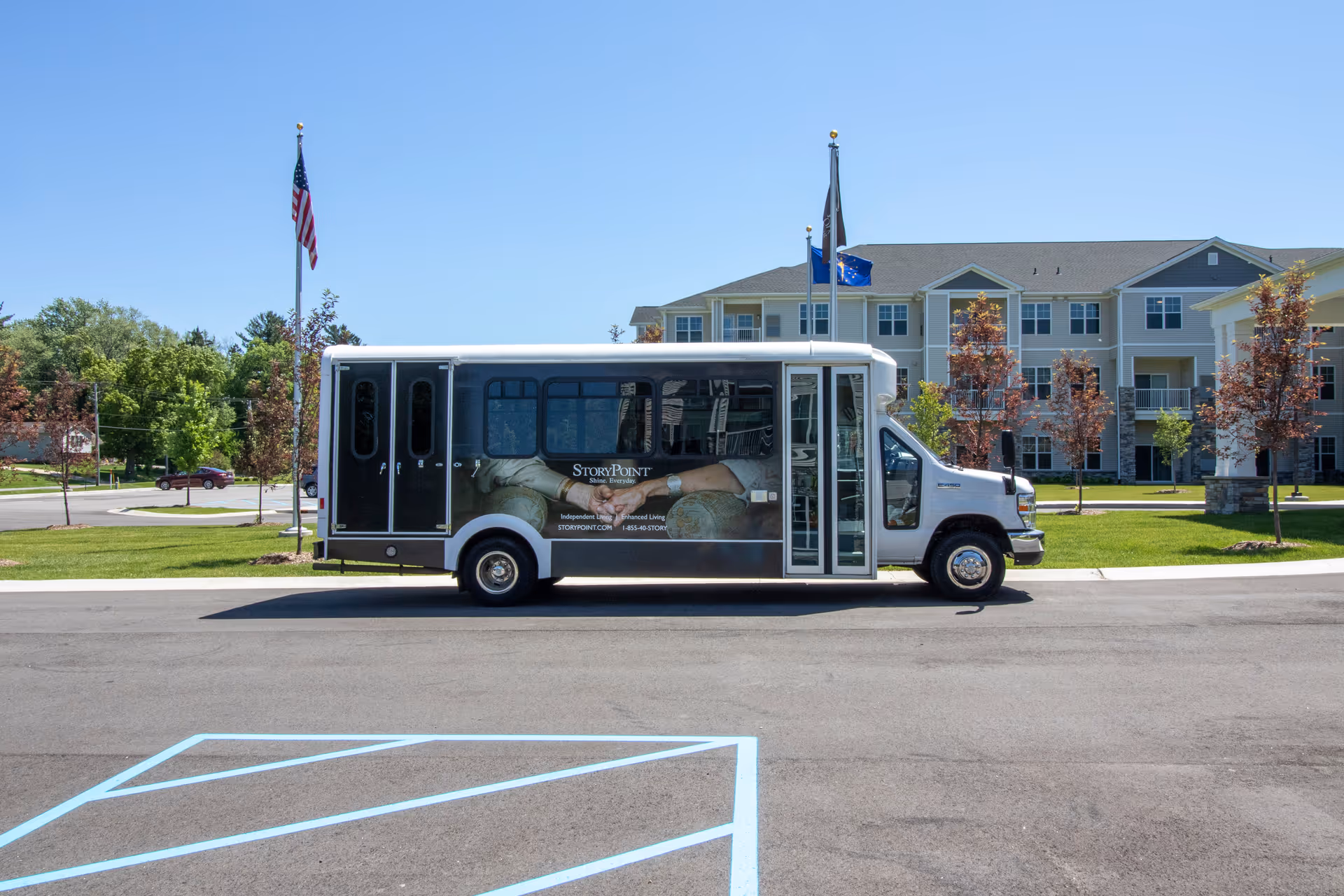 A StoryPoint Chesterfield shuttle bus parked on a road in front of a multi-story senior living facility building with green lawns, trees, and flagpoles displaying the American flag and other flags under a clear blue sky.