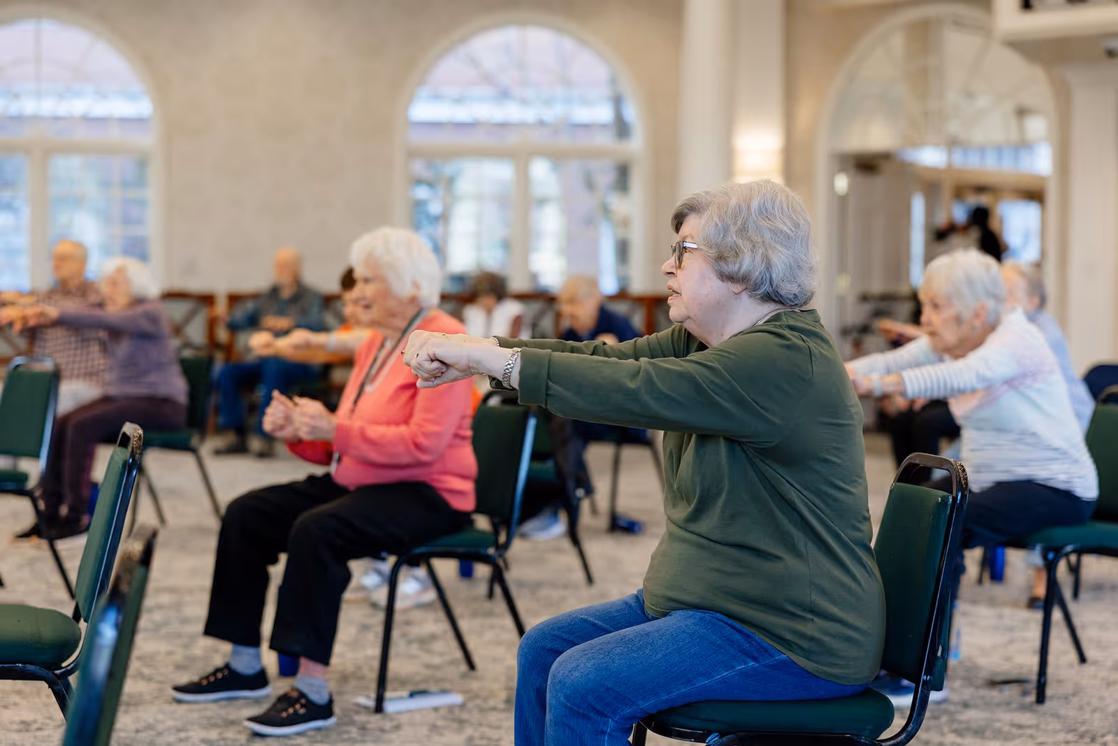 A group of elderly people sitting on chairs in a spacious room with large arched windows, participating in a seated exercise class by extending their arms forward.