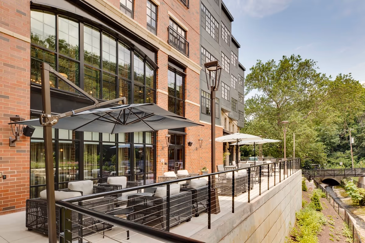 Outdoor patio area at The Residence at Bala Cynwyd featuring modern wicker seating with cushions, large umbrellas for shade, black metal railings, and a view of a small canal and greenery alongside the building.