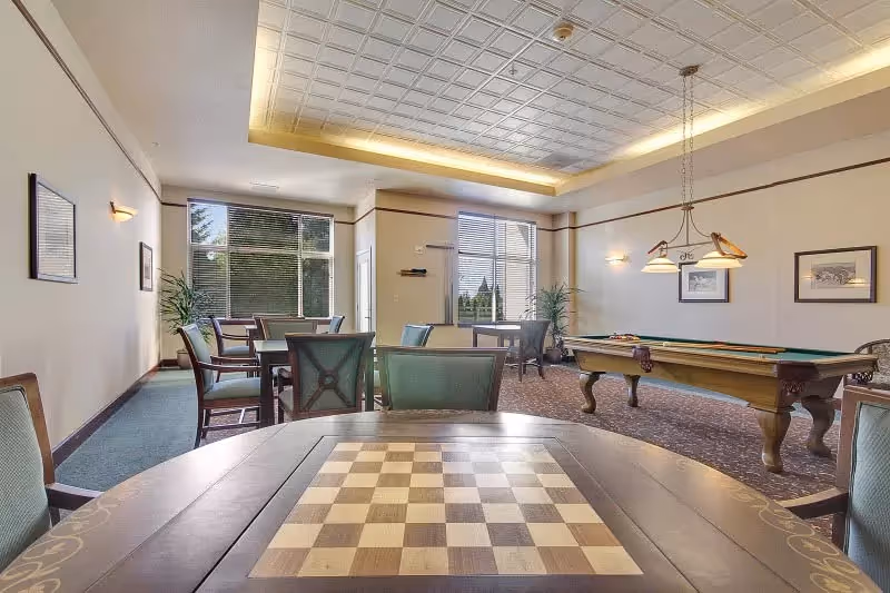 A well-lit game room with a chessboard table in the foreground, several chairs and tables arranged around the room, a pool table with overhead lights, framed artwork on the walls, large windows letting in natural light, and potted plants near the windows.