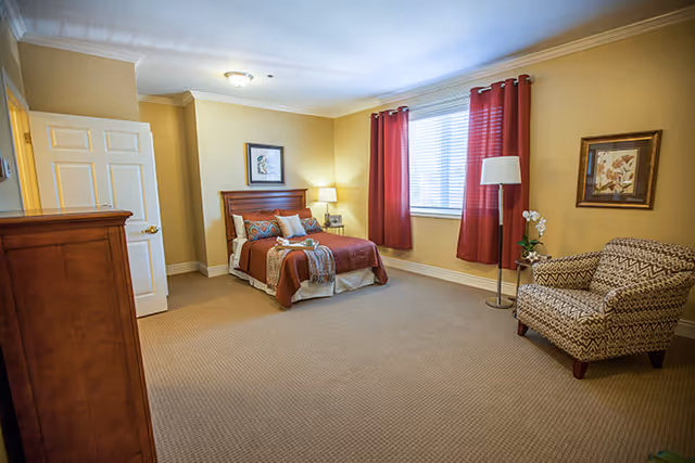 A spacious furnished bedroom with a bed, nightstand, dresser, armchair, and a window with red curtains.