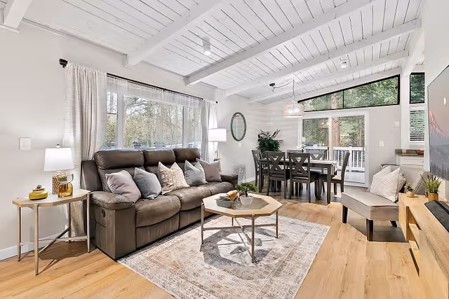 Bright open-concept living and dining area with a sofa, coffee table, dining table, and large windows under a white-beamed ceiling.