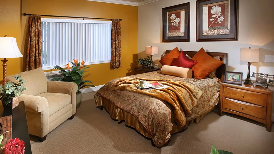 A cozy bedroom with a large bed covered in patterned bedding and multiple pillows in shades of orange and red. A soft brown blanket is draped over the bed with a tray holding a cup and a magazine. There are two framed floral artworks hanging above the bed. On either side of the bed are wooden nightstands with lamps, a framed photo, and a vintage telephone. To the left, there is a beige armchair next to a window with vertical blinds and patterned curtains. A green plant is placed near the window, and the walls are painted in warm tones of yellow and beige.