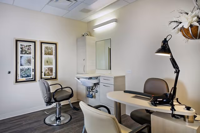 A small salon or grooming room with a salon chair, a sink with a mirror, a desk with a lamp, and two framed pictures on the wall. The room has light-colored walls and dark wood flooring.
