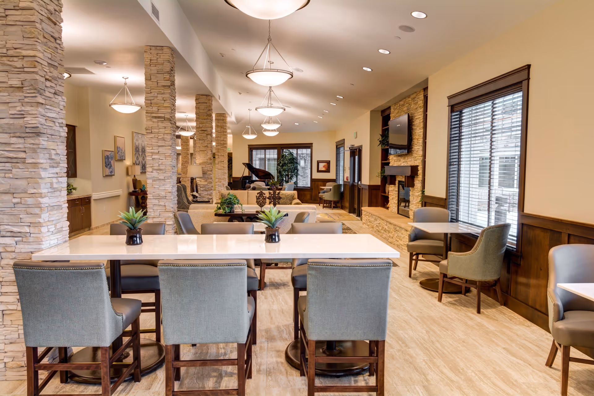 A spacious and well-lit common area in an assisted living facility featuring a long white table with six gray upholstered chairs, stone pillars, multiple hanging light fixtures, a fireplace with a mounted TV, large windows with blinds, and comfortable seating areas including sofas and armchairs. There is also a grand piano in the background and decorative plants on the tables.