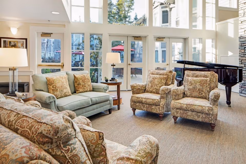 A bright and spacious living room area with large windows and glass doors letting in natural light. The room is furnished with a light green sofa, two patterned armchairs, and a patterned couch. There are two side tables with lamps, and a black grand piano is positioned near the stone wall on the right. Outside the glass doors, there are red umbrellas and outdoor seating visible.