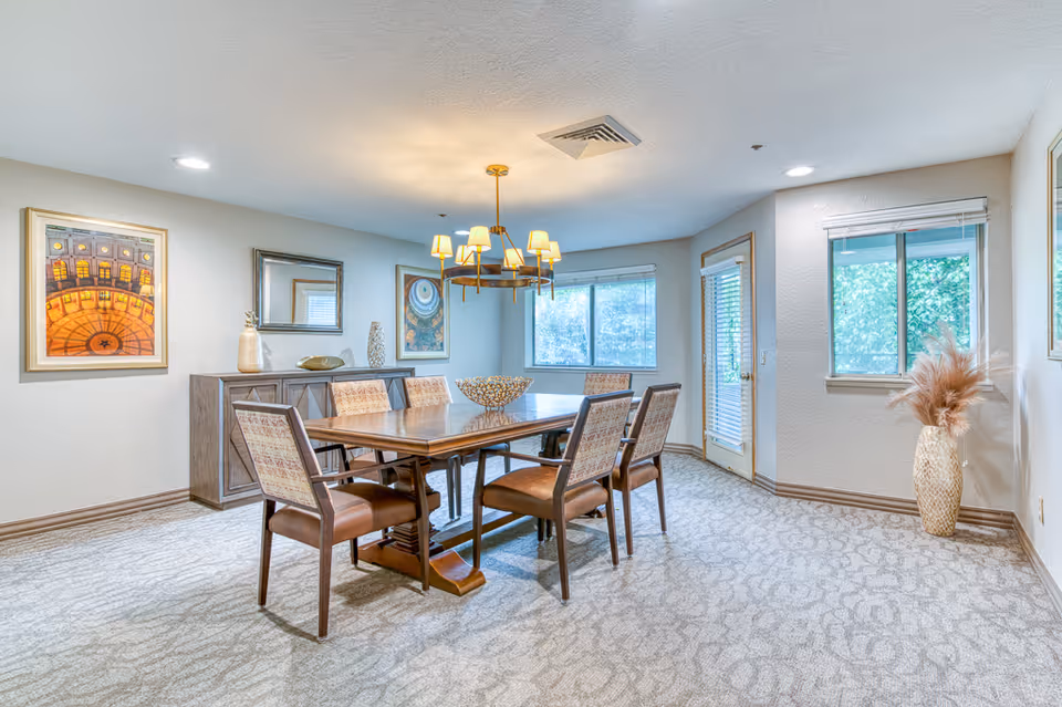 A bright dining room with a wooden table and six upholstered chairs. The room features a modern chandelier hanging above the table, a sideboard with decorative items, framed artwork on the walls, carpeted floor, and windows letting in natural light. There is also a door with blinds and a large vase with dried plants in the corner.