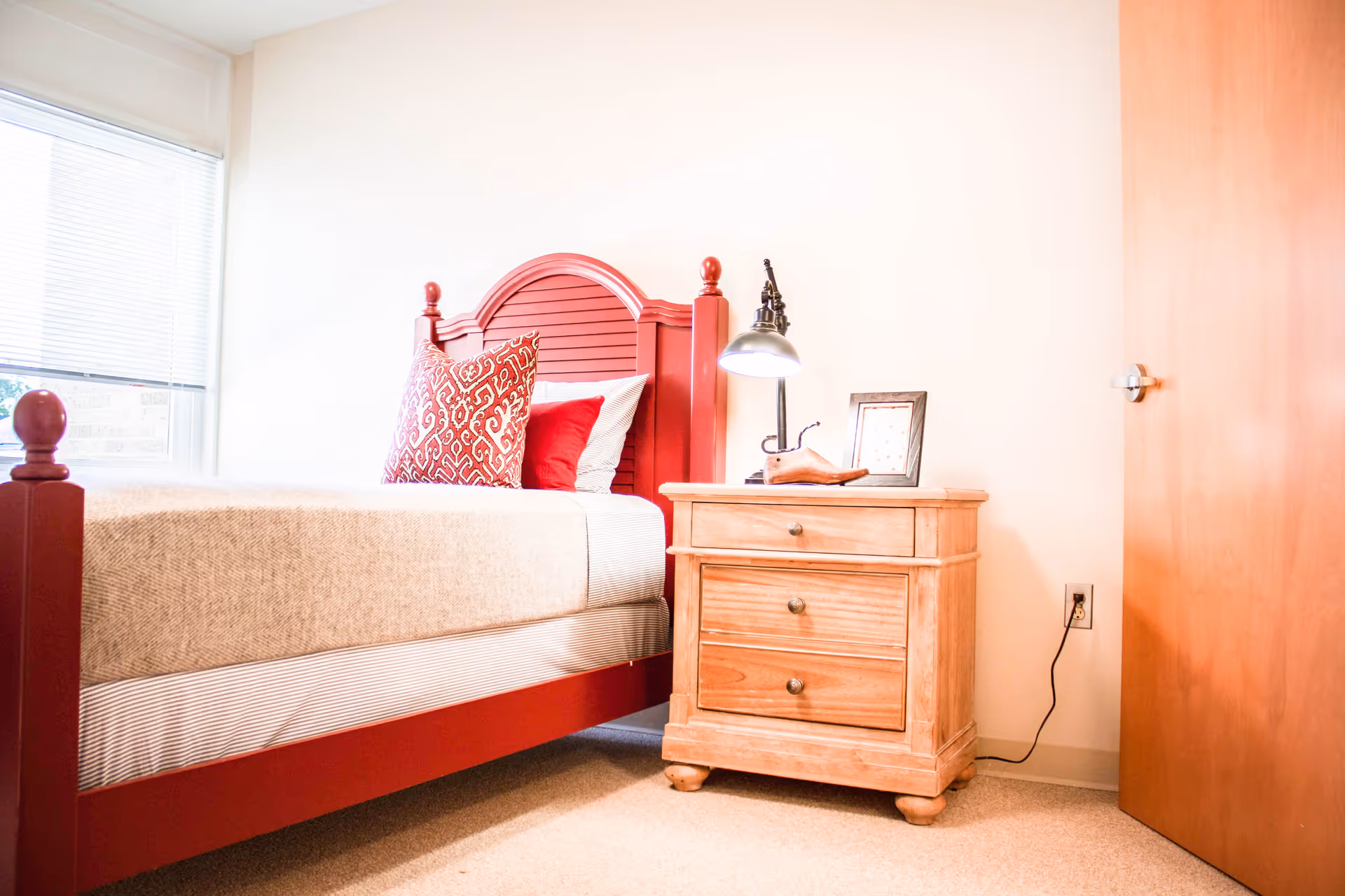 A bright bedroom with a red wooden bed frame, beige bedding, and red and patterned pillows. Next to the bed is a wooden nightstand with three drawers, a desk lamp, a framed picture, and a decorative shoe. A window with blinds is on the left side, and a wooden door is on the right.