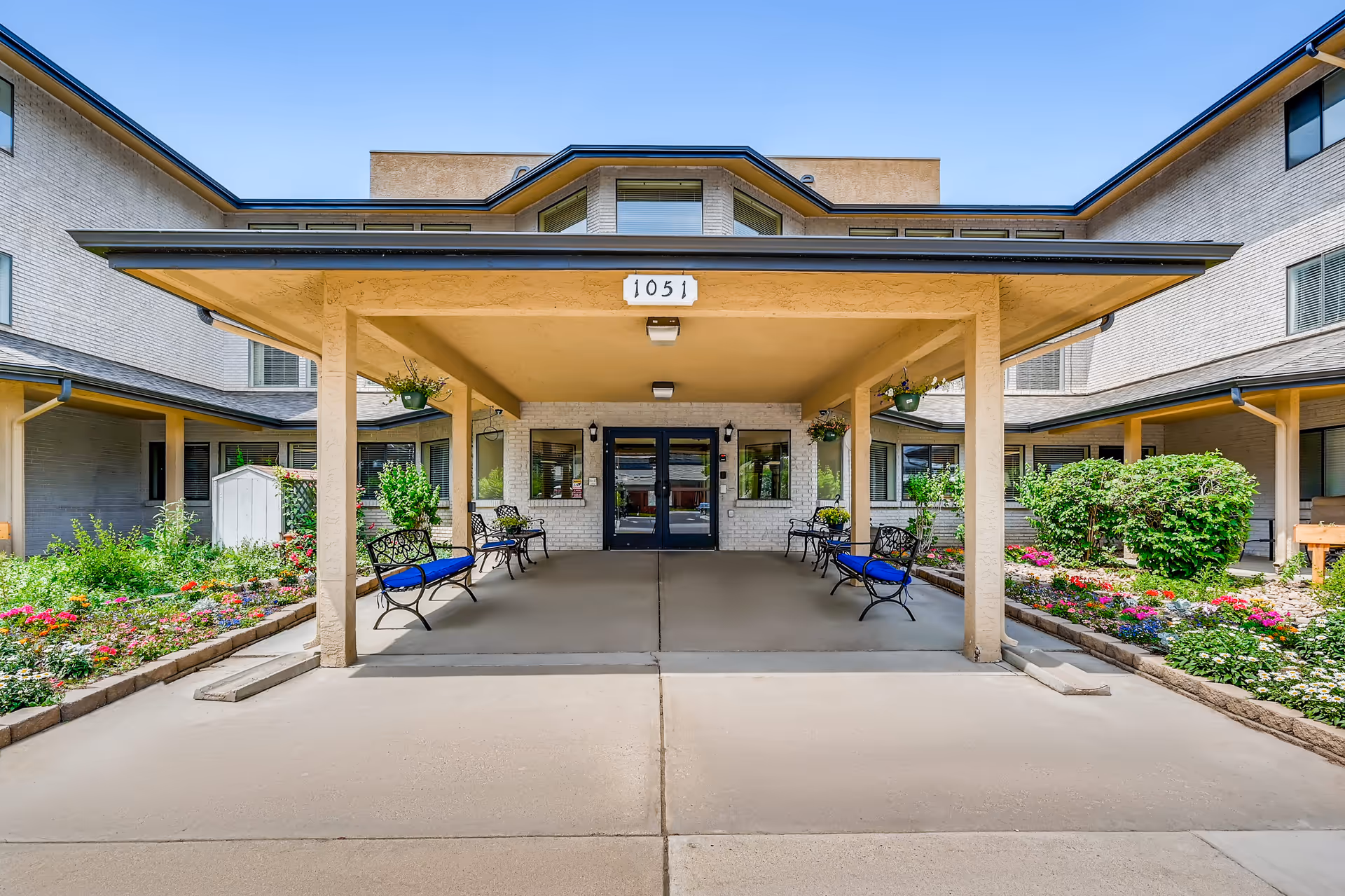 Front entrance of a senior living facility with a covered drop-off area, benches with blue cushions, hanging flower pots, and colorful flower beds on either side. The building is light-colored with multiple windows and the number 1051 displayed above the entrance.